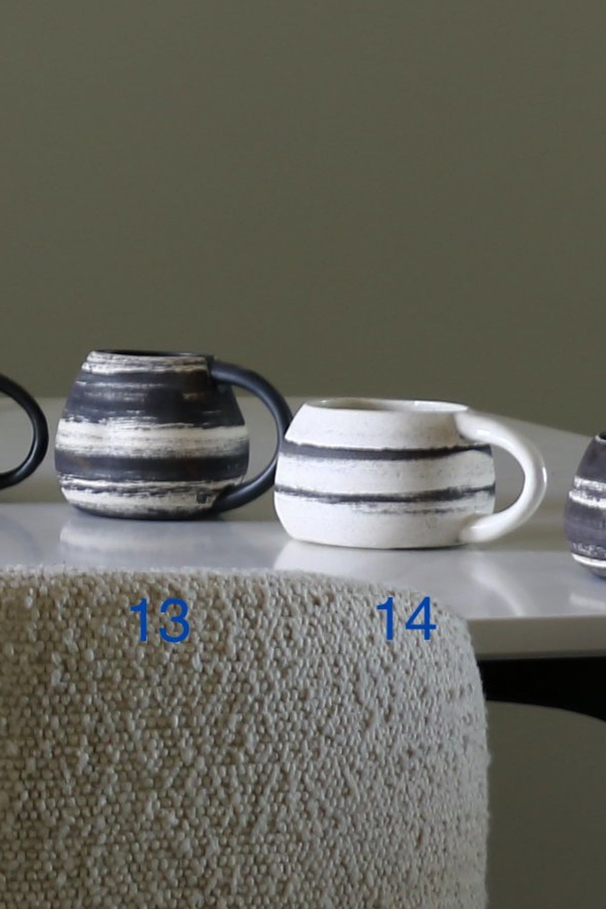 Two ceramic mugs with a rustic hand-painted finish, one dark gray and one white, both featuring horizontal black stripes and dark handles, displayed on a white table with a textured beige cloth draped over the front. Blue numbers '13' and '14' are overlaid near the base of the mugs.