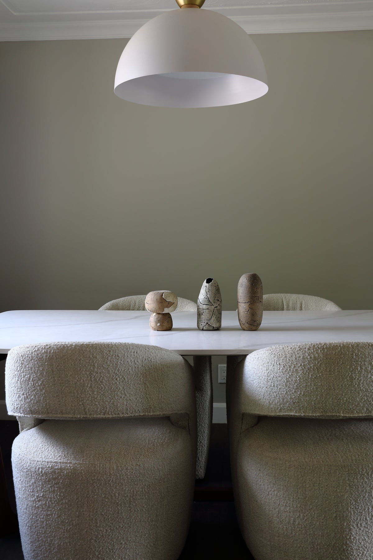 A dining room scene with a white table and four upholstered chairs in a warm beige bouclé fabric, under a large white dome pendant light. Three small decorative vases sit on the table against a muted beige-gray wall.