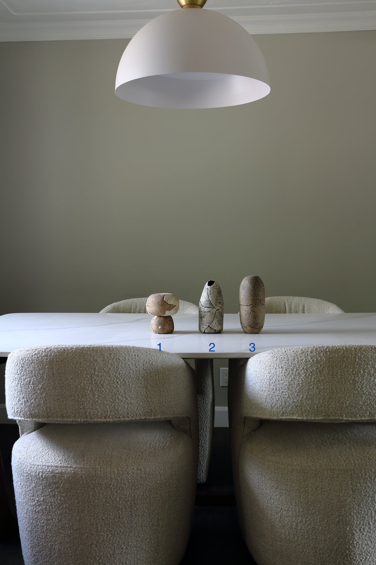A dining table with three upholstered chairs in a neutral beige setting, featuring three small decorative vases on top and a large white dome pendant light hanging above.