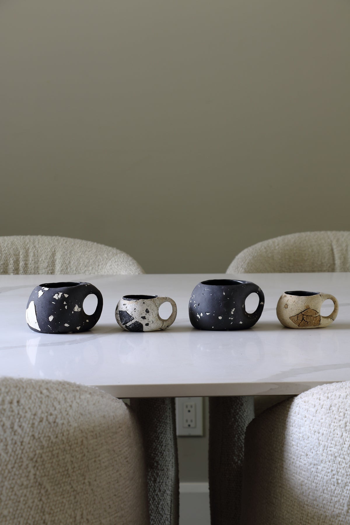 Four ceramic mugs with unique organic shapes and handles are lined up on a white marble dining table surrounded by textured beige boucle chairs.