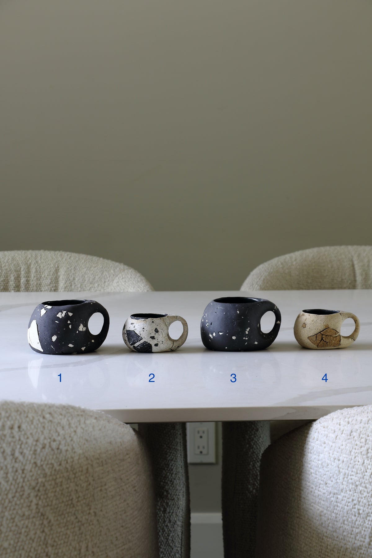 Four ceramic mugs with unique patterns and colors arranged on a white marble table in a dining room setting, numbered 1 to 4 below them.