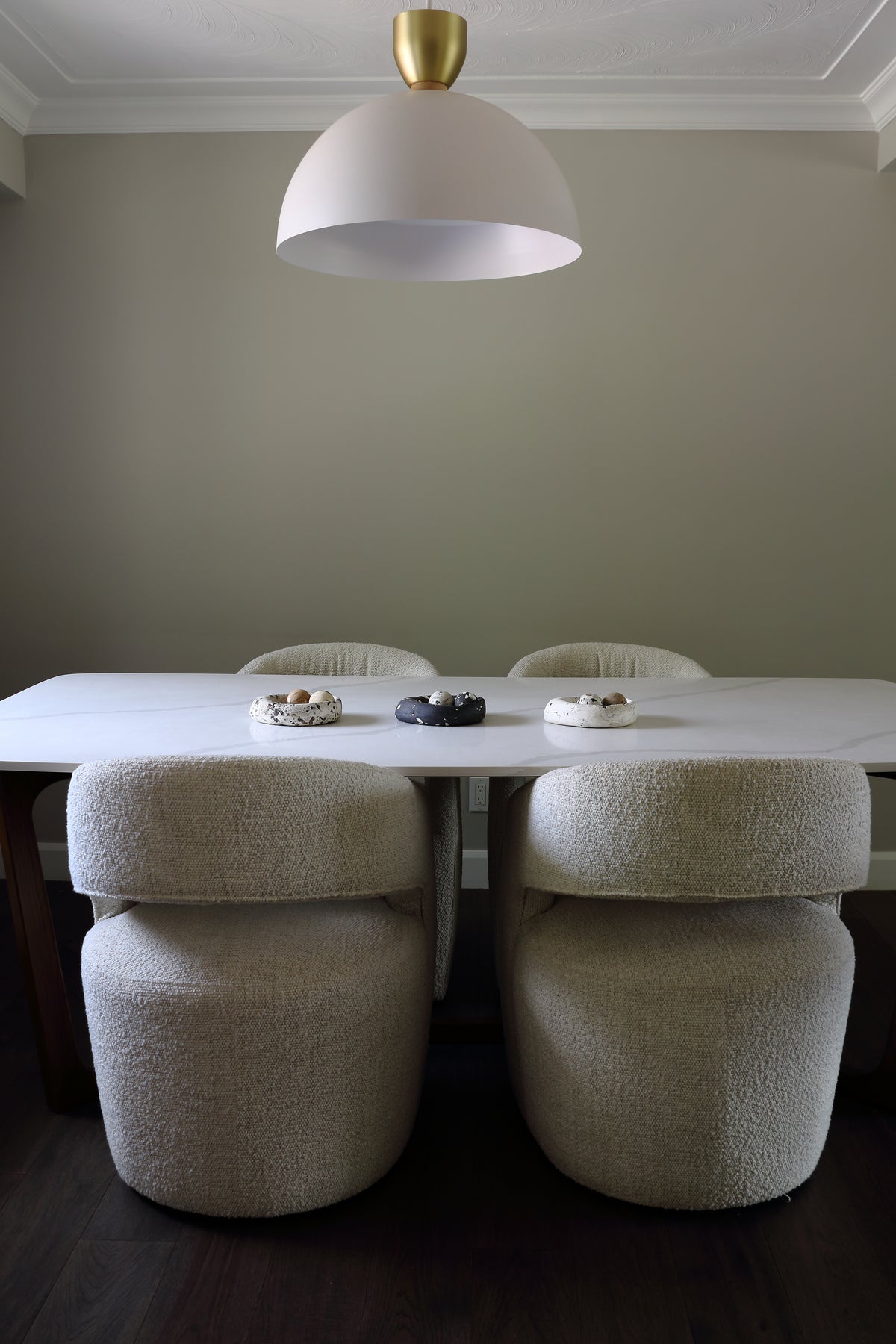 A modern dining room setup featuring a white rectangular table, four upholstered bouclé dining chairs in a light beige color, and a large white dome pendant light with a gold-toned top cap hanging above.