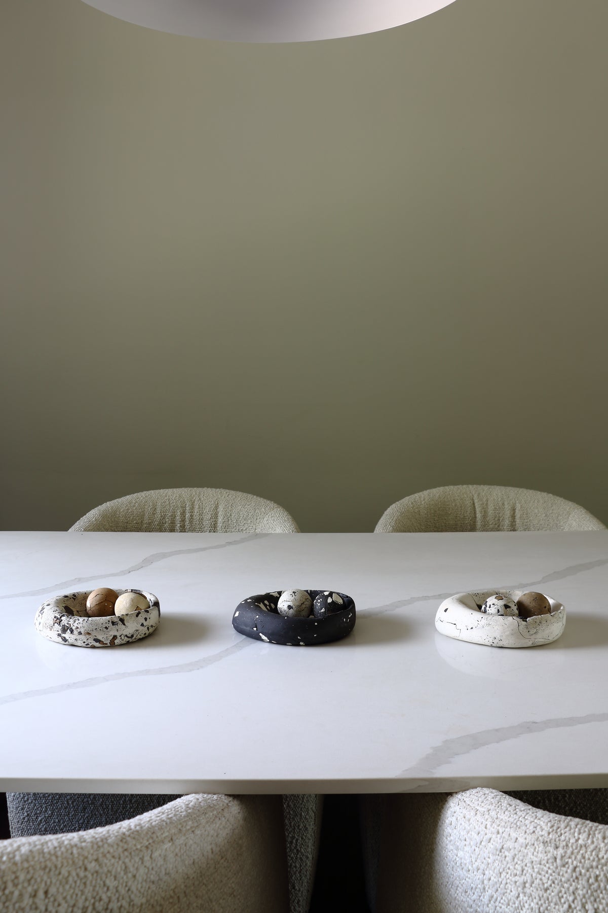 Three small decorative egg-shaped trays or bowls arranged on a white marble dining table, with upholstered chairs and a muted sage-green wall in the background.