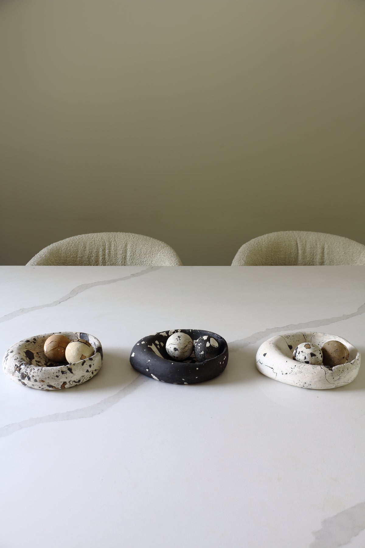Three small ceramic egg-shaped bowls with speckled textures, containing quail eggs, arranged on a white marble table with two chairs in the background against a beige wall.