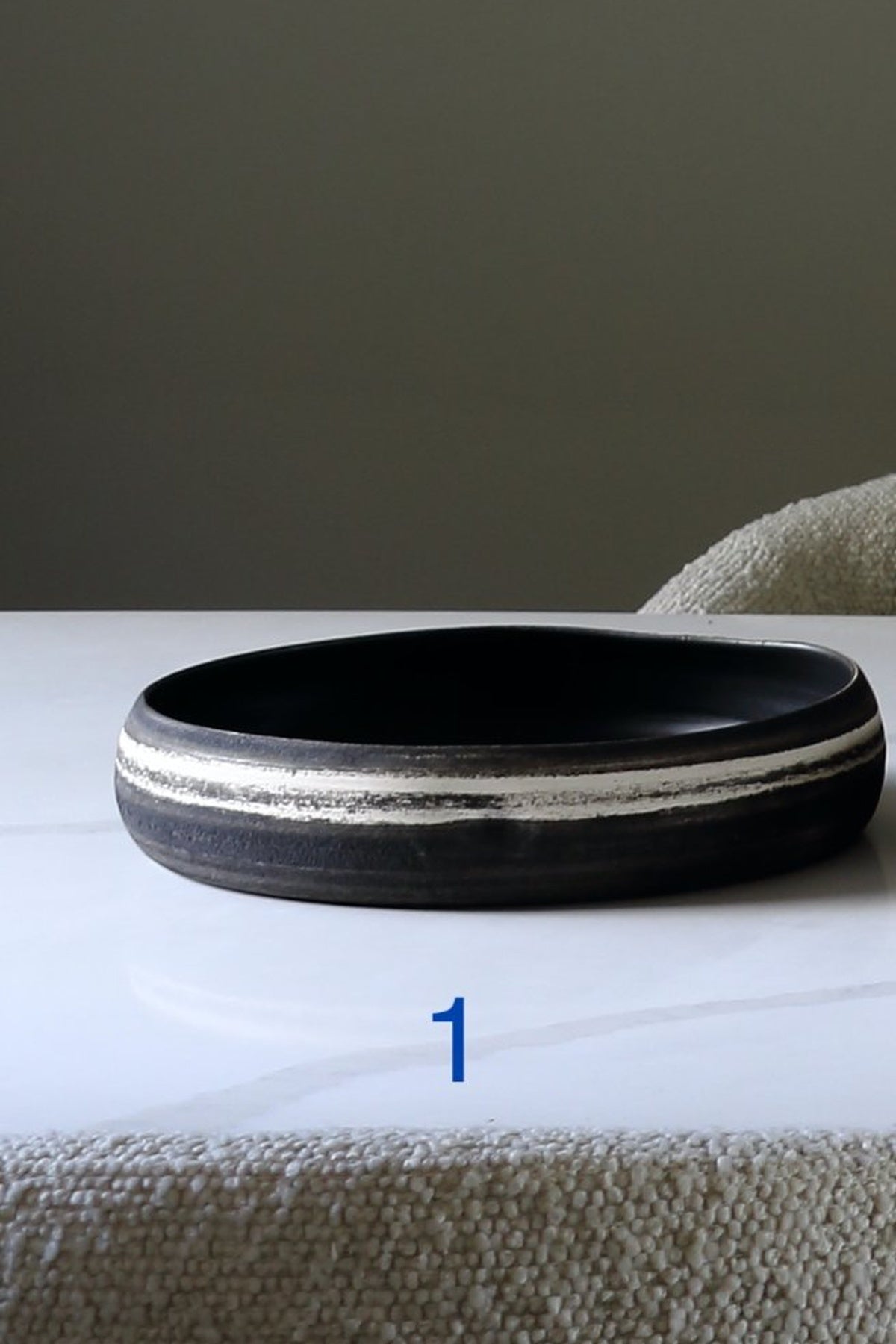 A wide, shallow black ceramic bowl with two textured silver bands, resting on a white marble surface with a textured beige fabric in the foreground and background.