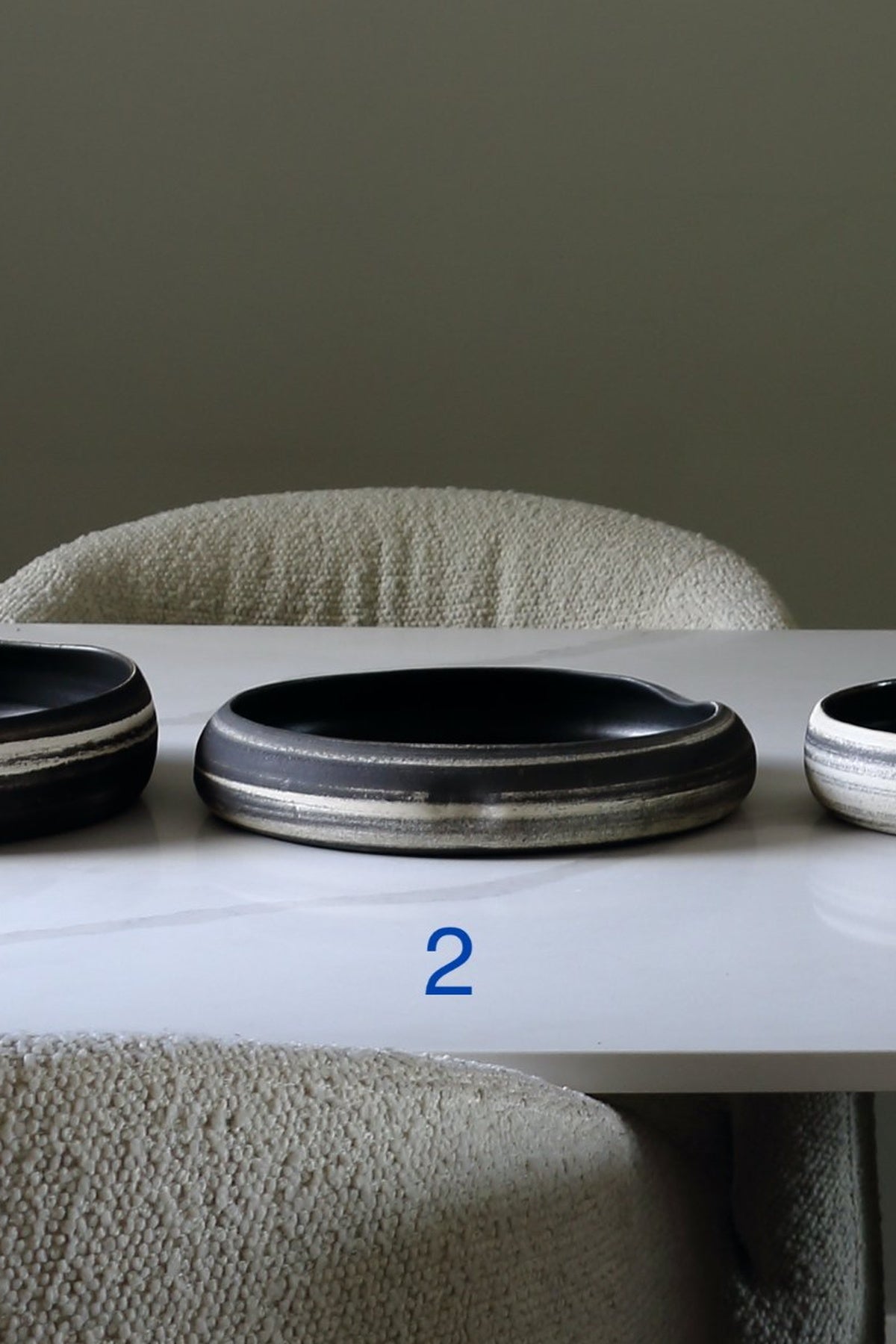 A dark ceramic oval tray or shallow bowl with a metallic silver rim, displayed on a white table in front of a textured beige chair. A blue number '2' is overlaid near the bottom.