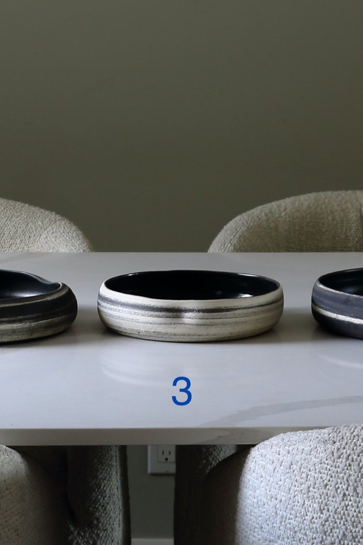 A ceramic bowl with a wide silver-toned band and a glossy black interior, displayed on a white table between upholstered chairs. A blue number 3 is printed below the bowl.