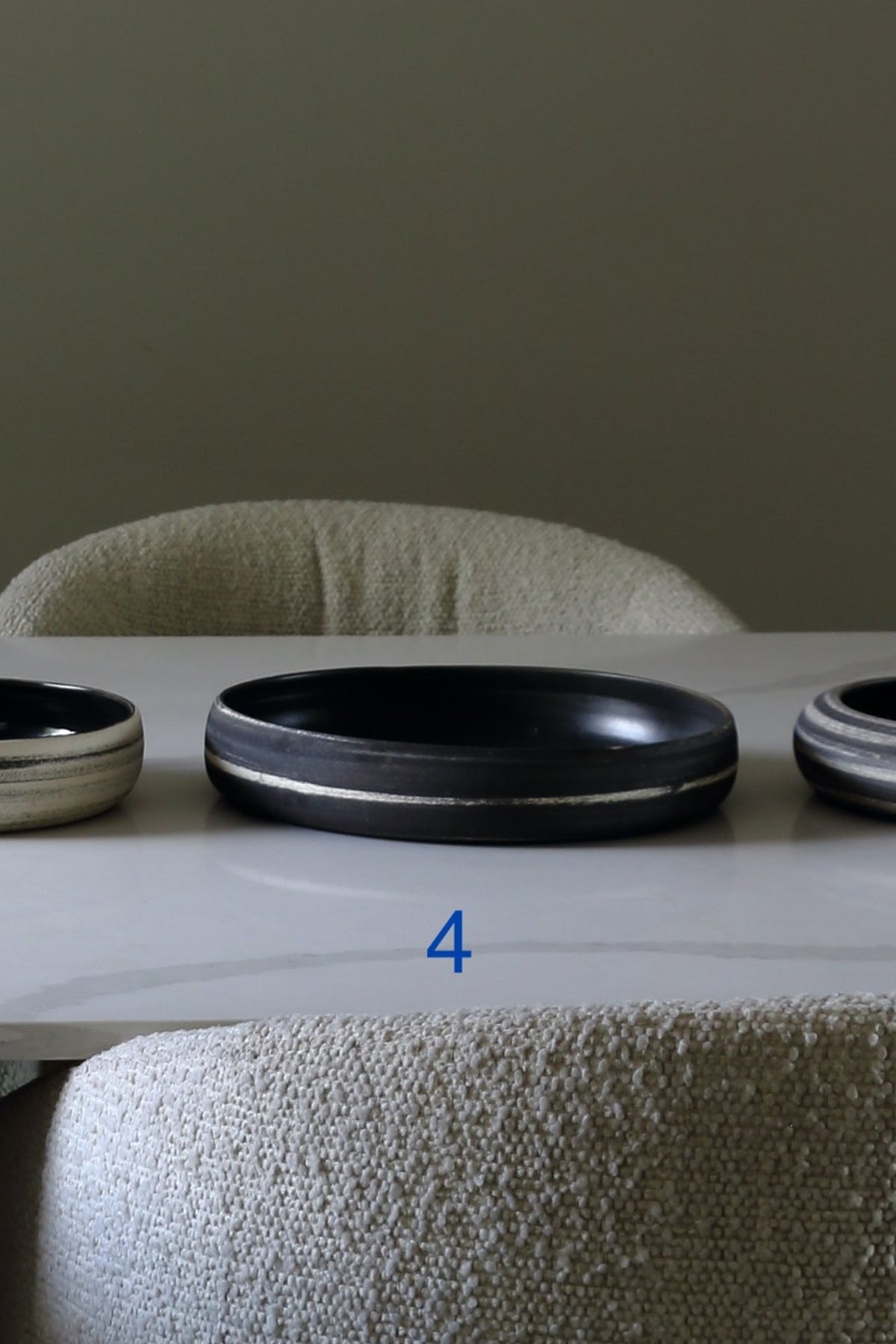 A black oval ceramic tray with a thin silver metallic rim, displayed on a white marble table in front of a textured beige upholstered chair. A blue number '4' is overlaid near the bottom.