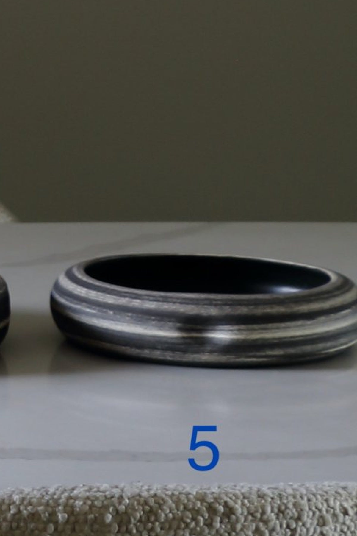 A close-up of a wide bangle bracelet with a textured, layered silver and black striped pattern, resting on a light-colored surface with a gray background.