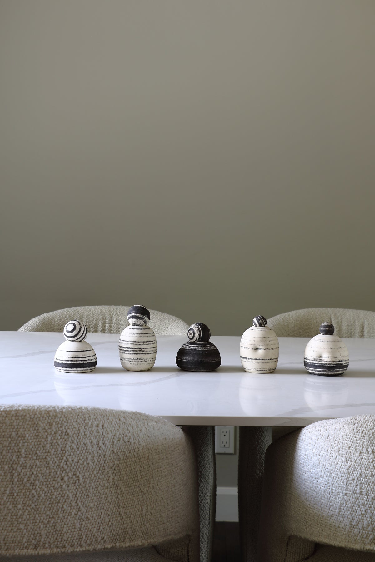 Five small decorative ceramic bird figurines in black, white, and charcoal are lined up on a white marble dining table between upholstered beige chairs against a muted sage green wall.