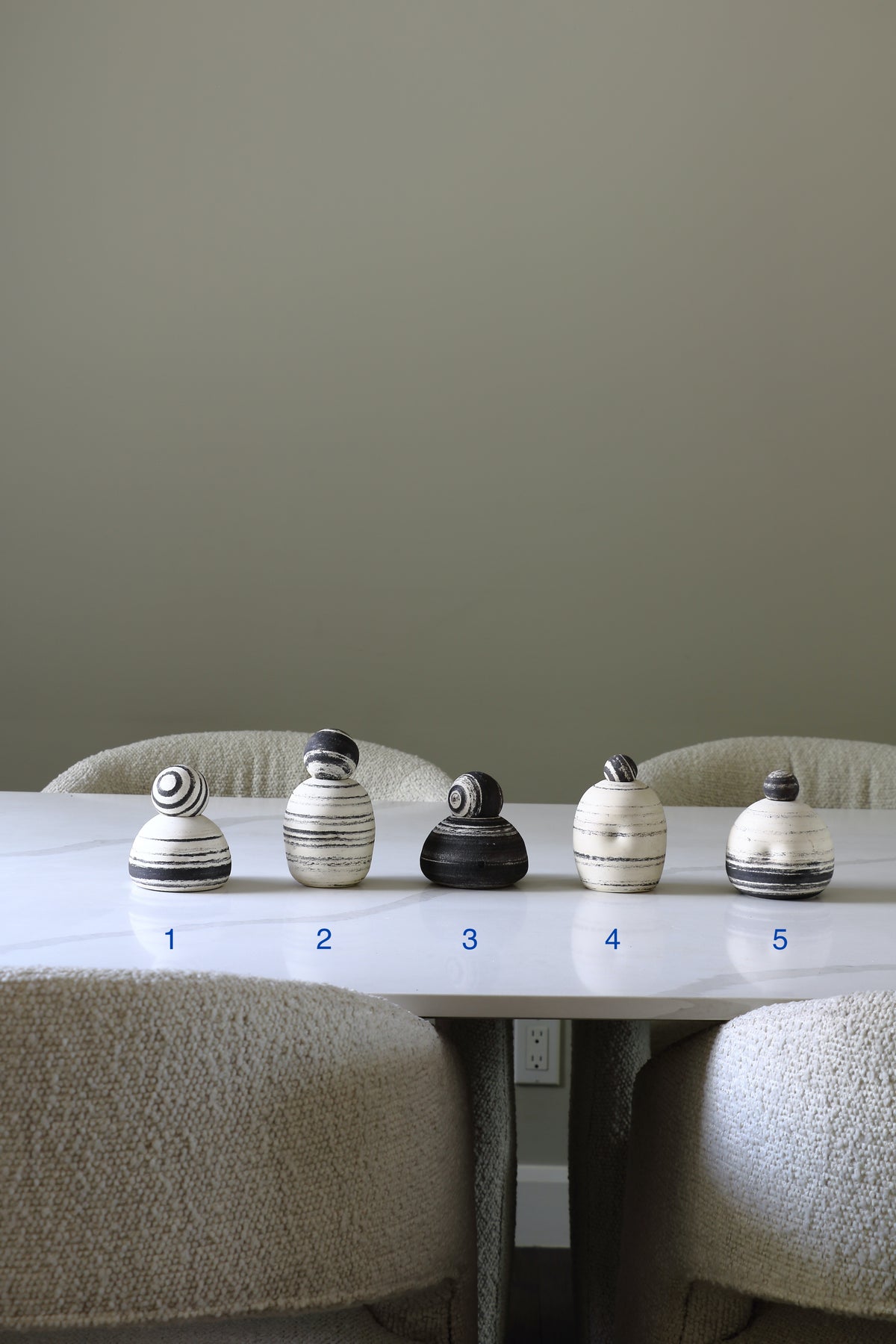 Five small decorative ceramic vases or bottles with matching lids are lined up on a white table in a dining setting. The vases have black and white marbled or striped patterns, with two in dark charcoal/black and three in off-white/cream.