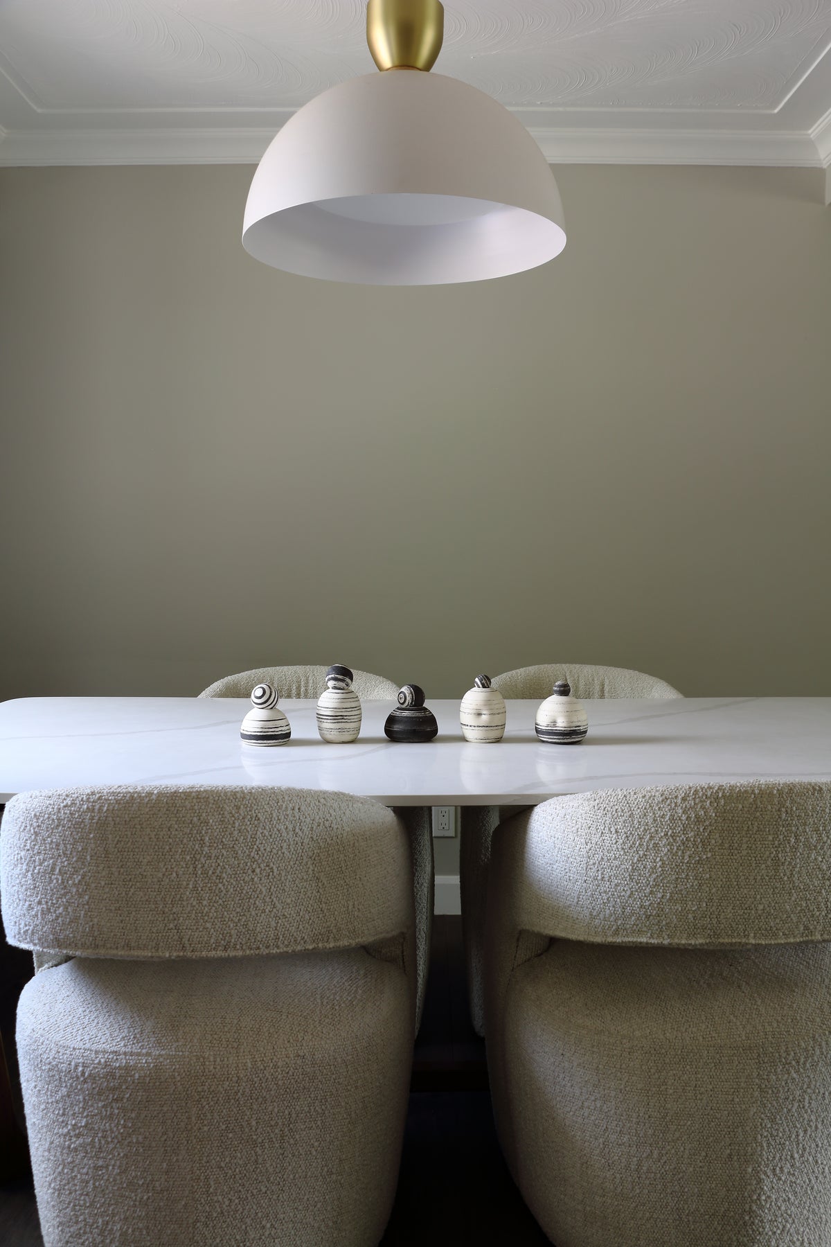 A modern dining room scene featuring a white table with four bouclé upholstered dining chairs, a white dome pendant light with a gold finial, and a neutral gray wall.