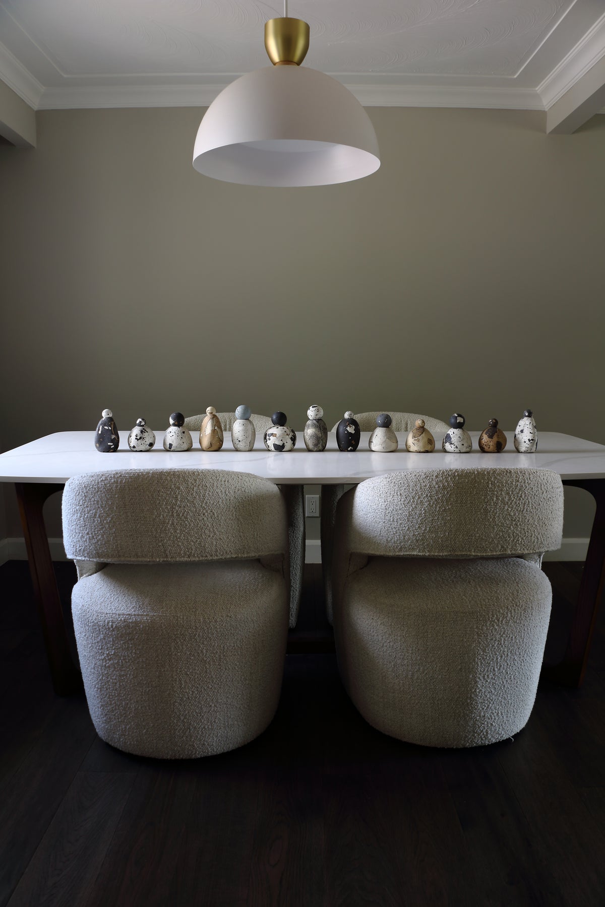 A dining area featuring a white rectangular table with two upholstered chairs in a textured cream bouclé fabric, set against a warm gray wall under a white dome pendant light with a gold top.