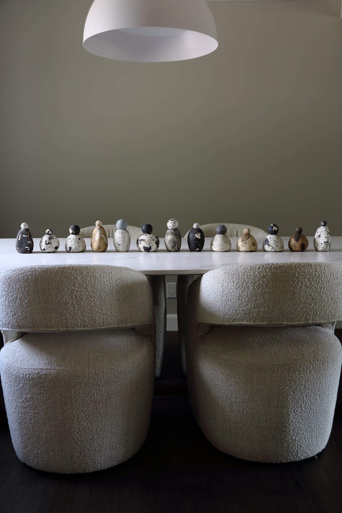 Two modern bouclé barrel chairs in a light beige textured fabric, positioned on either side of a white dining table. A row of small decorative ceramic figurines sits on the tabletop, and a white dome pendant light hangs from above against a neutral gray wall.