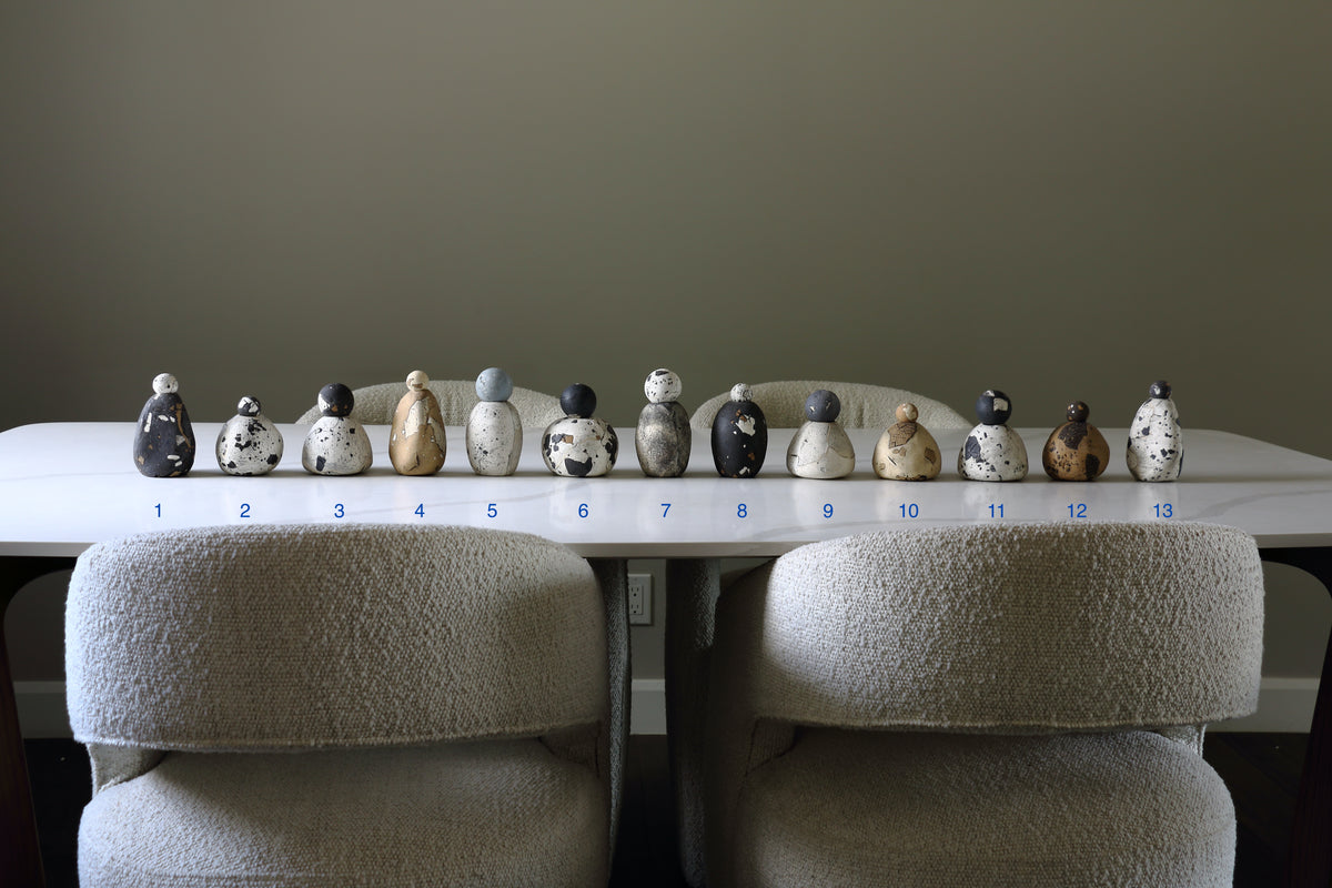 A row of 13 small hand-painted ceramic Russian dolls arranged on a white table, with two beige boucle chairs in front and blue numbers 1 through 13 printed along the table edge.
