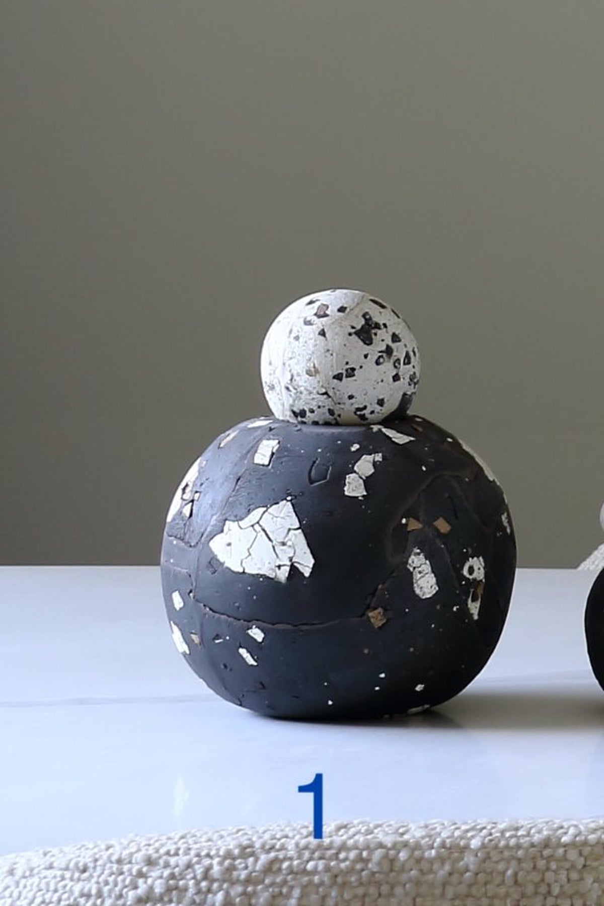 A decorative sculptural object with a large black spherical base, a smaller white speckled sphere on top, and irregular white and metallic flecks throughout, displayed on a white surface against a plain gray background.