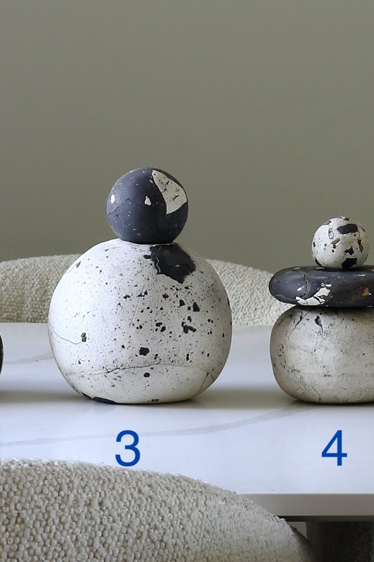 Two decorative stacked stone sculptures on a white table against a neutral gray background. The left sculpture is labeled with the blue number 3, and the right one with the blue number 4.