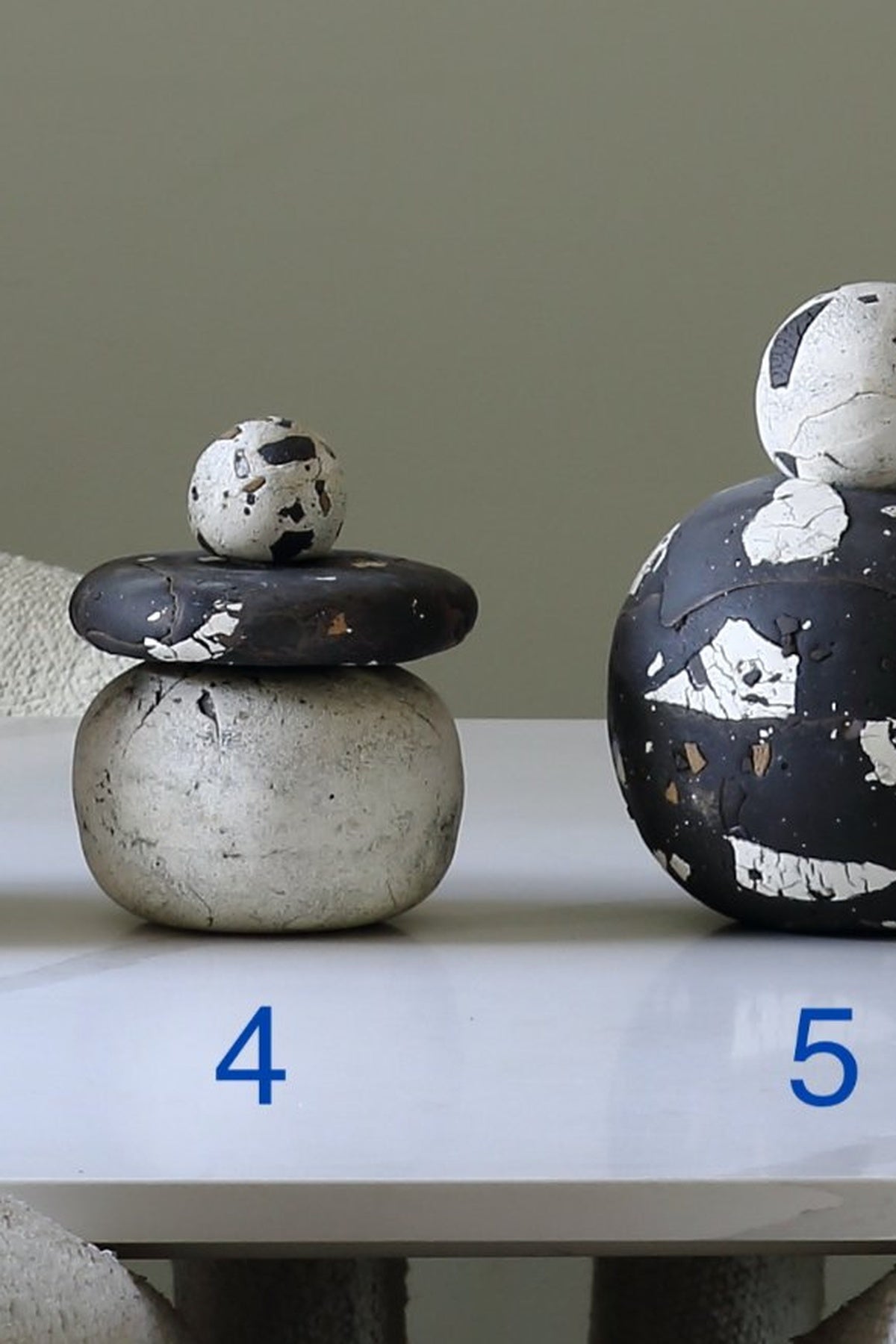 Two decorative stacked ceramic objects displayed on a white surface against a muted gray-green background. The left object is labeled 4 and consists of a white rounded base with a dark charcoal lid and a small white top sphere with dark speckles.