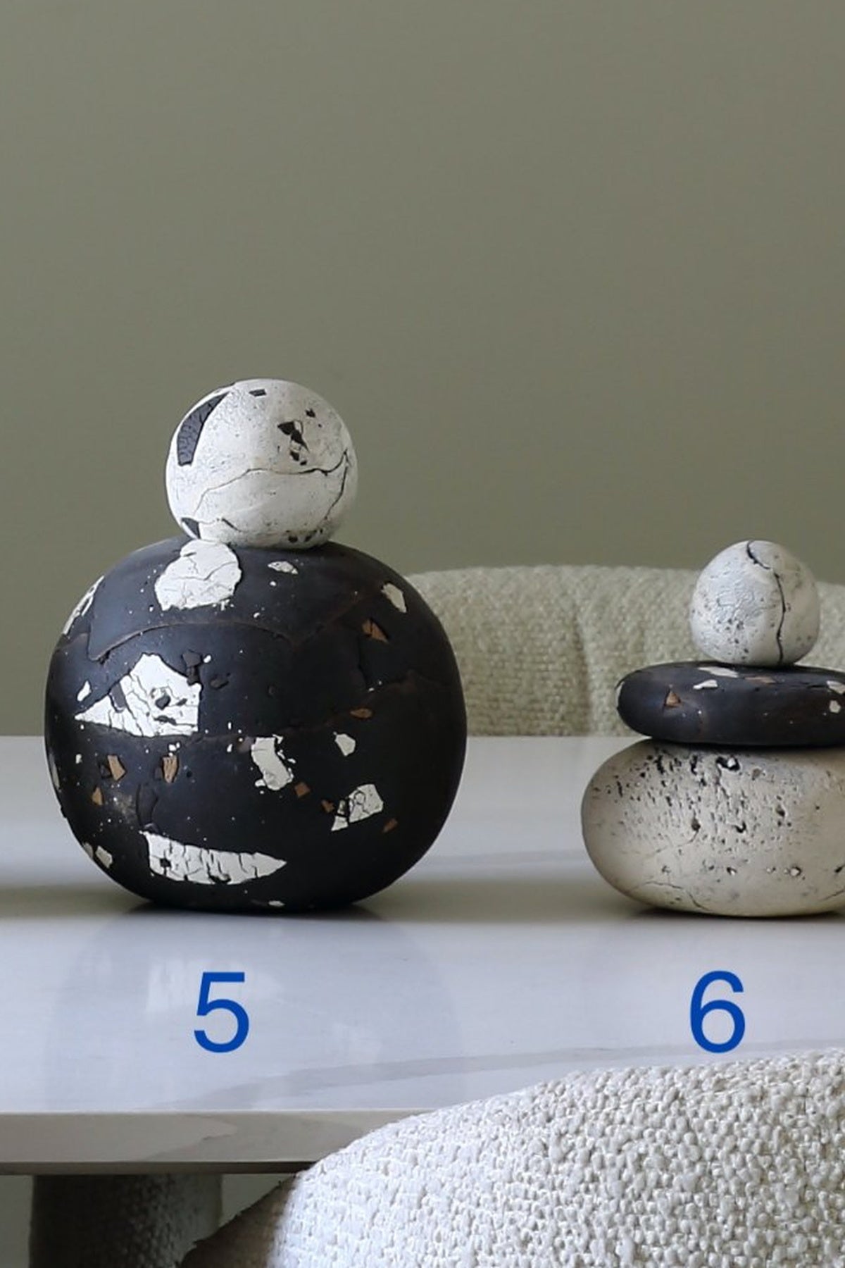 Two decorative stacked stone-like sculptures displayed on a white table against a muted green wall. The left sculpture is larger and matte black with white distressed patches and a white rounded top; the right sculpture is smaller, with a dark top, a speckled off-white middle layer, and a light cream base.