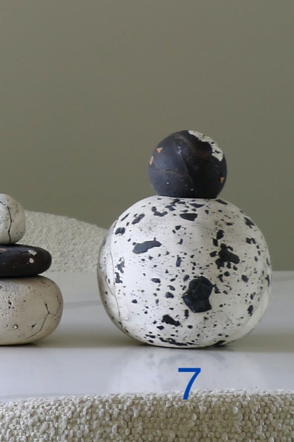 A decorative ceramic or stone-like object with a white rounded body covered in black speckles and a dark charcoal spherical topper, displayed on a white surface against a muted beige background. A blue number 7 appears near the bottom.