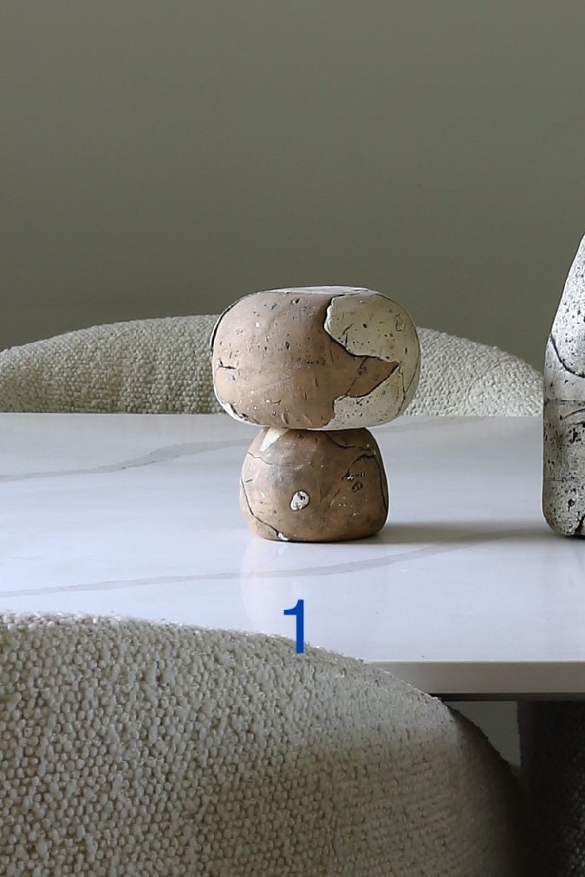 A small rustic wooden mushroom-shaped object with a cracked, weathered finish sits on a white tabletop in a styled interior setting with textured beige chairs and a neutral wall.