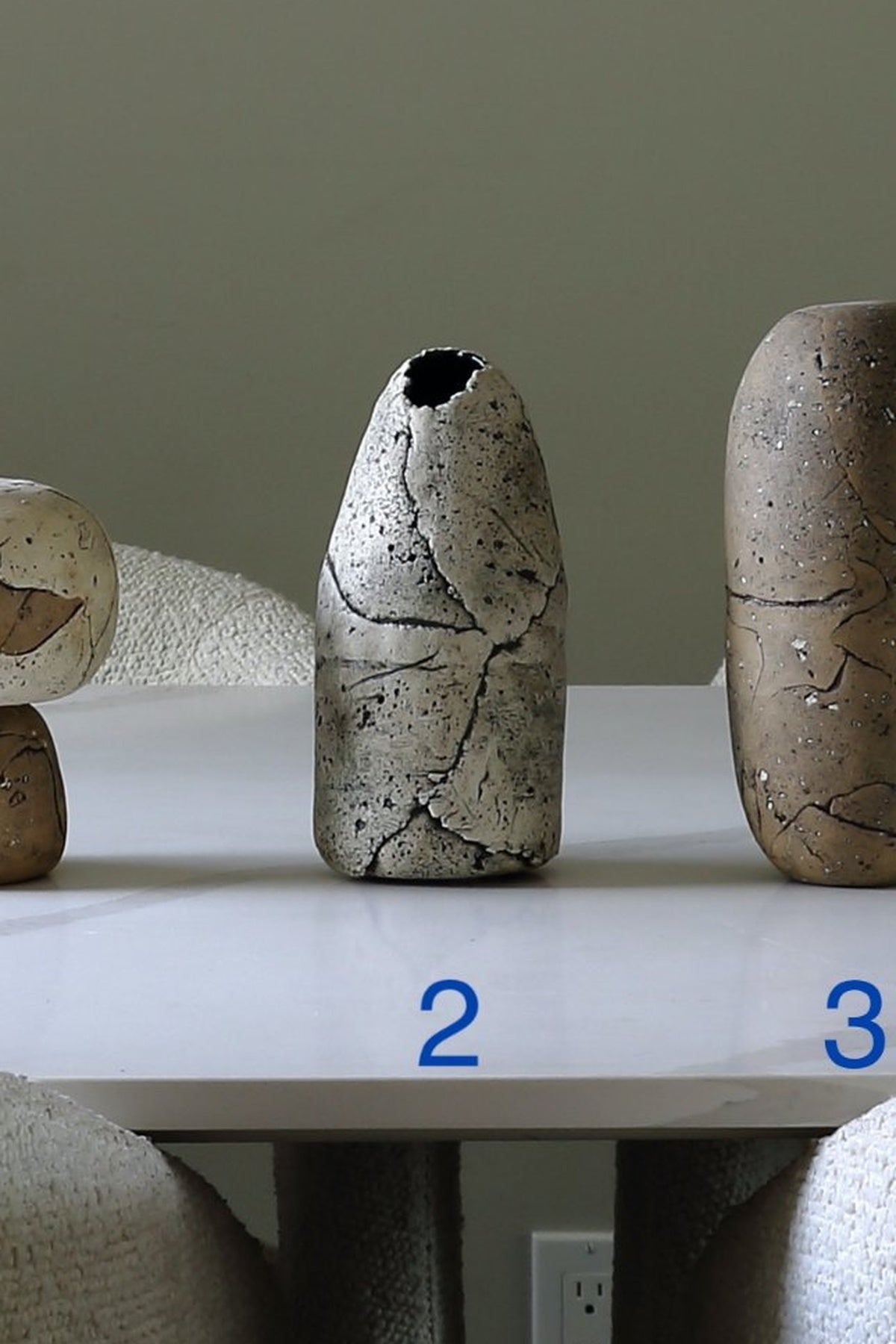 Two textured ceramic vases displayed on a white table against a muted green-gray wall. The left vase is partially cropped, and the right vase is fully visible.
