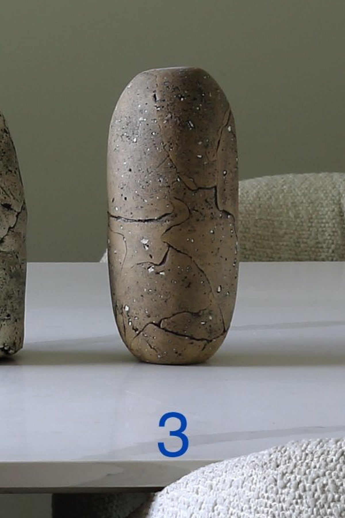 A small rounded ceramic vase with a textured, earthy brown and gray glaze and dark crackle detailing, standing on a white table. A blue number '3' is printed below the vase.