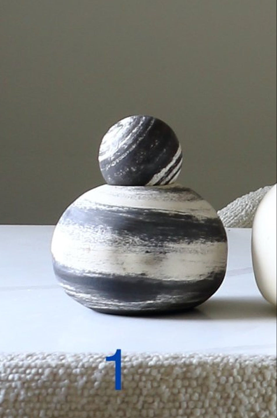 A small decorative sculpture with a round black-and-white marbled top sphere resting on a larger oval base with horizontal black and off-white marbling, displayed on a white surface against a neutral gray wall.