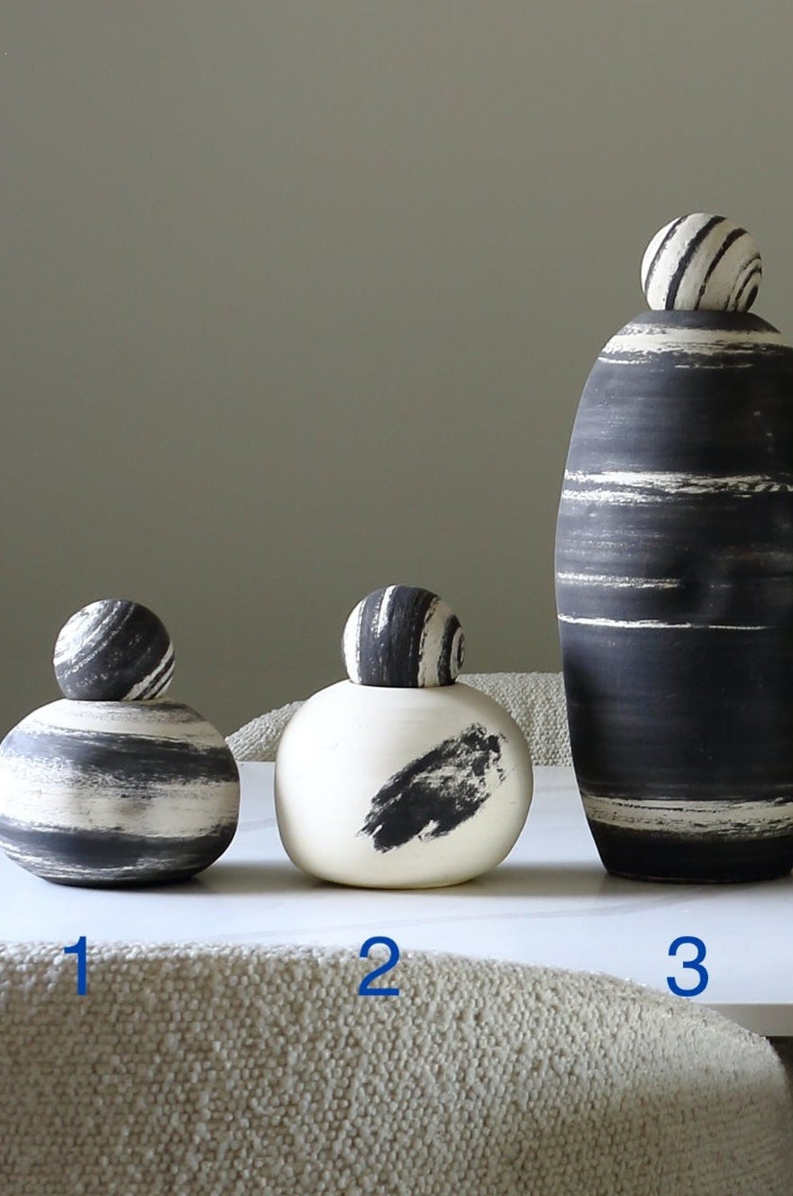 Three hand-painted ceramic duck figurines in black, white, and gray brushstroke patterns, displayed on a white table in front of a textured beige chair against a neutral gray wall. The numbers 1, 2, and 3 are printed in blue near the respective vases.