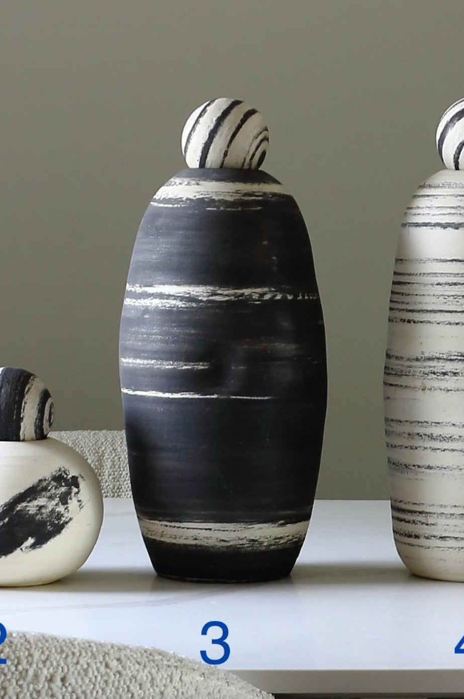 Three decorative ceramic vases with matching lids are displayed on a white table against a neutral gray wall. The vases are numbered 2, 3, and 4 in blue text.