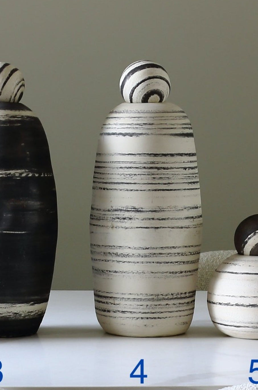 Three decorative ceramic lidded vases with striped patterns in black and off-white, displayed on a white table with blue numbers 3, 4, and 5 underneath them.