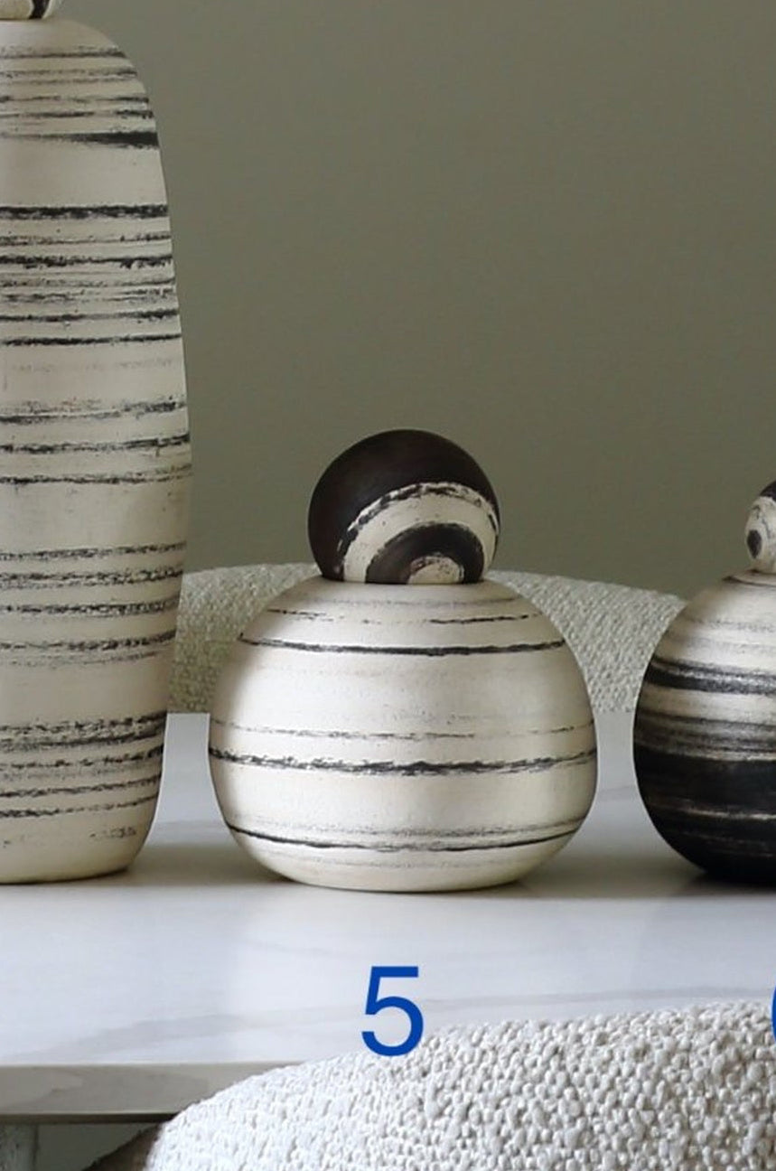 Three decorative ceramic vases with lids in a horizontal striped pattern, displayed on a white shelf with blue numbers 4, 5, and 6 underneath them.