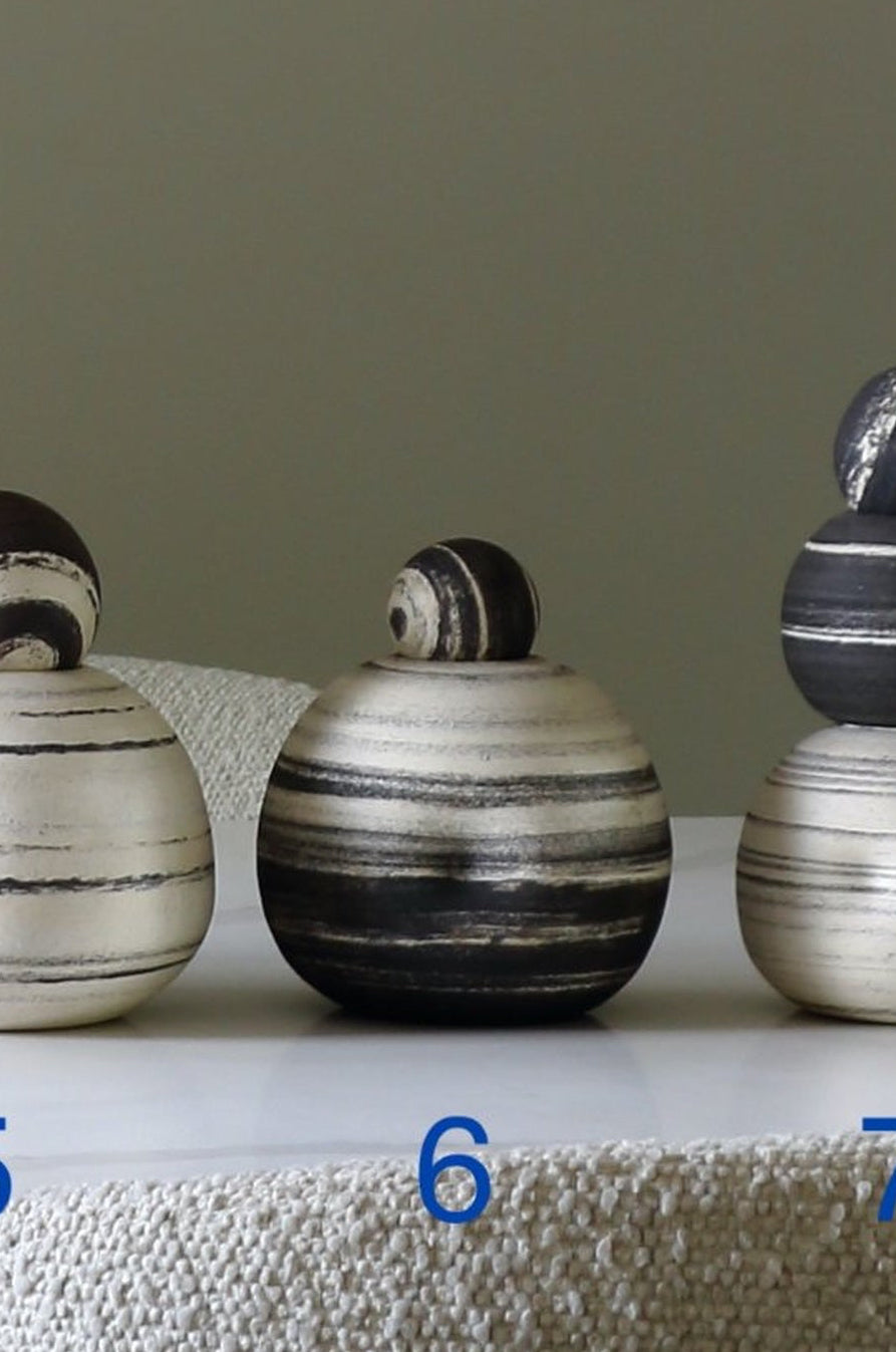 Three stacked ceramic decorative vases with a striped pattern in black, white, and gray, displayed on a textured white surface against a neutral gray background. Blue numbers 5, 6, and 7 are overlaid below each vase.