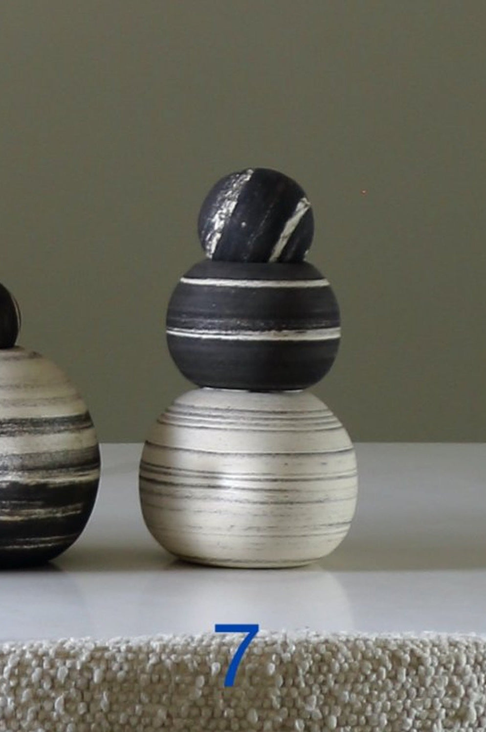Two stacked ceramic decorative objects with horizontal marbled stripes in black, white, and gray, displayed on a white surface against a muted gray-green background. Blue numbers 6 and 7 are overlaid near the base.