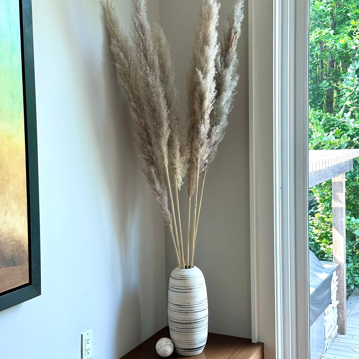 A tall decorative vase filled with dried pampas grass, placed on a wooden table in a corner near a window with a view of green trees outside.
